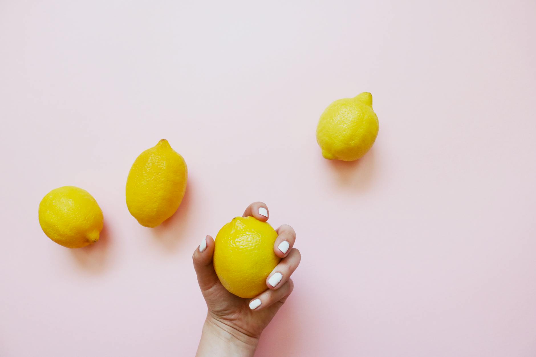 A hand holding a fresh lemon against a soft pink background surrounded by additional lemons